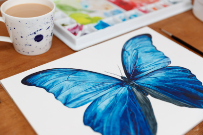 A detailed, brightly coloured watercolour painting of a blue Giant Blue Morpho (Morpho didius) on a wooden table. In the background is a paint pallet covered in paints and a cup of tea in a white mug which is decorated with blue splashes.