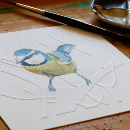A watercolour painting of a Blue Tit (Cyanistes caeruleus). The blue & yellow bird rests on blind embossed branches. The photograph is angled highlighting the embossing. The painting is on a wooden table next to a paint brush & a small paint pallet.