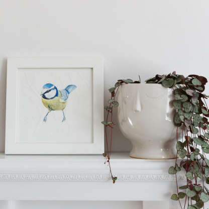 A watercolour painting of a Blue Tit (Cyanistes caeruleus). The blue & yellow bird rests on blind embossed branches. The painting is in a white wooden frame which is placed on a white fireplace. Next to the bird is a plant in a white plant pot.