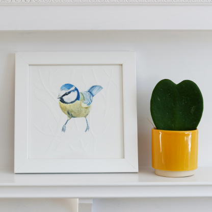 A watercolour painting of a Blue Tit (Cyanistes caeruleus). The painting is in a white wooden frame which is placed on a white fireplace. Next to the bird is green potted succulent in a small yellow pot.