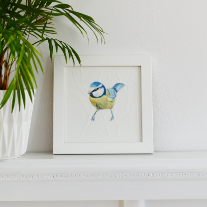 A watercolour painting of a Blue Tit (Cyanistes caeruleus). The blue & yellow bird rests on blind embossed branches. The painting is in a white wooden frame which is placed on a white fireplace. Next to the bird is green potted plant in a white pot.