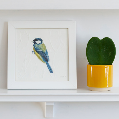 A watercolour painting of a Great Tit. The blue & yellow bird grasps onto blind embossed branches. The painting is in a white wooden frame which is placed on a white fireplace. Next to the bird is green potted succulent in a small yellow pot.