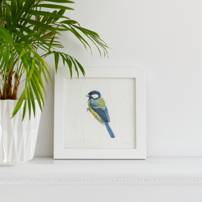 A watercolour painting of a Great Tit (Parus major). The blue & yellow bird grasps onto blind embossed branches. The painting is in a white wooden frame placed on a white fireplace. Next to the bird is green potted plant in a white pot.