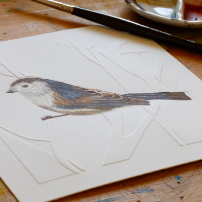 A watercolour painting of a Long Tailed Tit (Aegithalos caudatus). The brown & white bird grasps onto blind embossed branches. Angled to highlight the embossing, the painting lays on a wooden table next to a paint brush and a paint pallet.