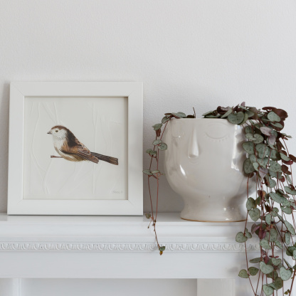 A watercolour painting of a Long Tailed Tit. The brown & white bird grasps onto blind embossed branches. The painting is in a white wooden frame which is placed on a white fireplace. Next to the bird is a string of hearts plant in a white plant pot.