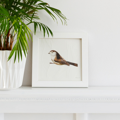 A watercolour painting of a Long Tailed Tit (Aegithalos caudatus). The brown & white bird grasps onto blind embossed branches. The painting is in a white wooden frame placed on a white fireplace. Next to the bird is green potted plant in a white pot.