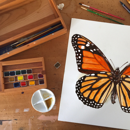 A detailed, brightly coloured watercolour painting of an orange Monarch Butterfly (Danaus plexippus) on a wooden table. On the table is a paint pallet covered in paints and paint brushes.