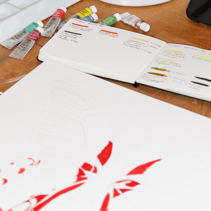 A process photograph of the watercolour painting of the red Scarlet Mormon butterfly. The painting has some red patches on it and it is on a wooden table top. In front of it is a book with colour swatches for the painting alongside paint tubes.